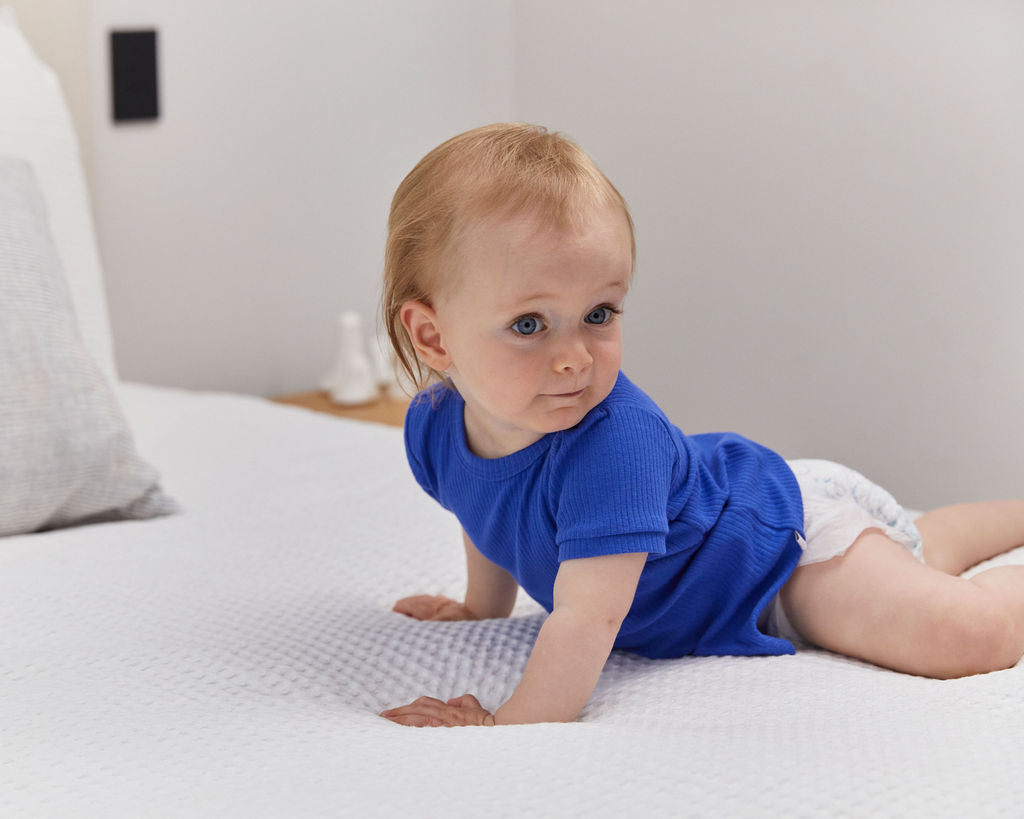 A baby in a blue top crawling across a white bed