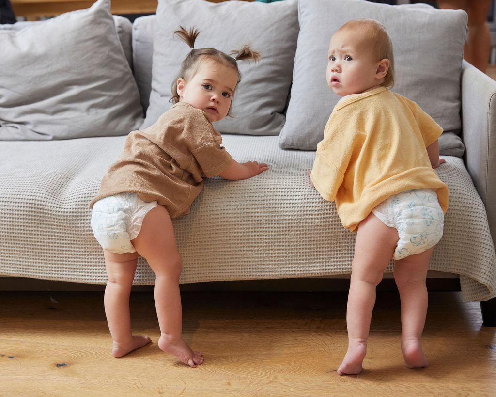 Two toddlers in Treasures nappies standing against a sofa