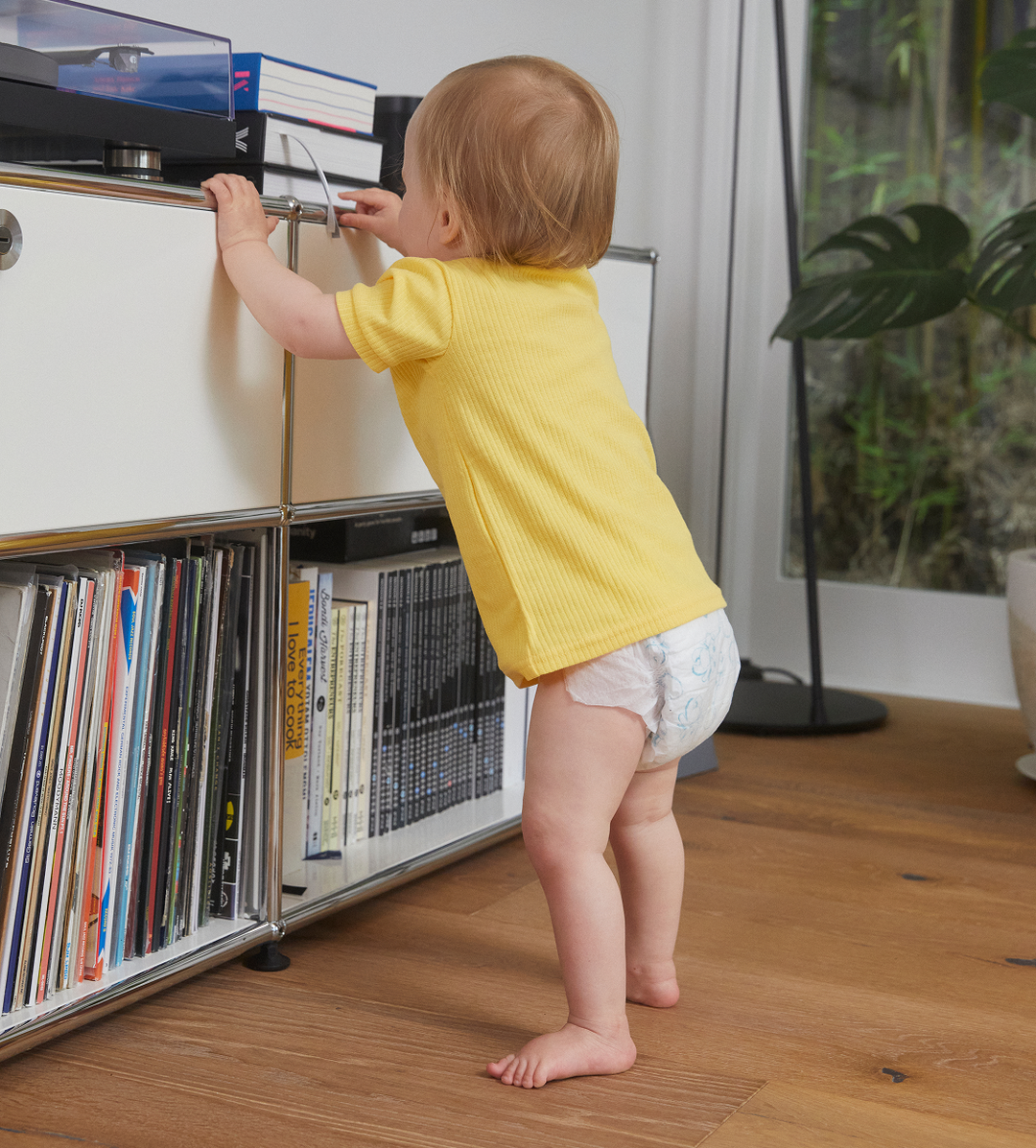 A toddler in a yellow top stands at a shelf, wearing a Treasures nappy